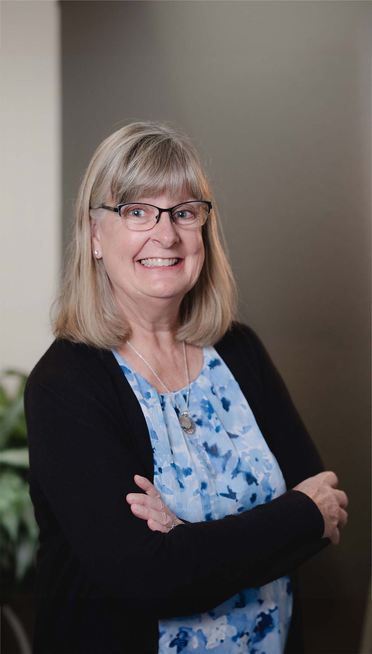 Smiling middle-aged woman with glasses, wearing a blue floral blouse and black cardigan, standing with arms crossed.