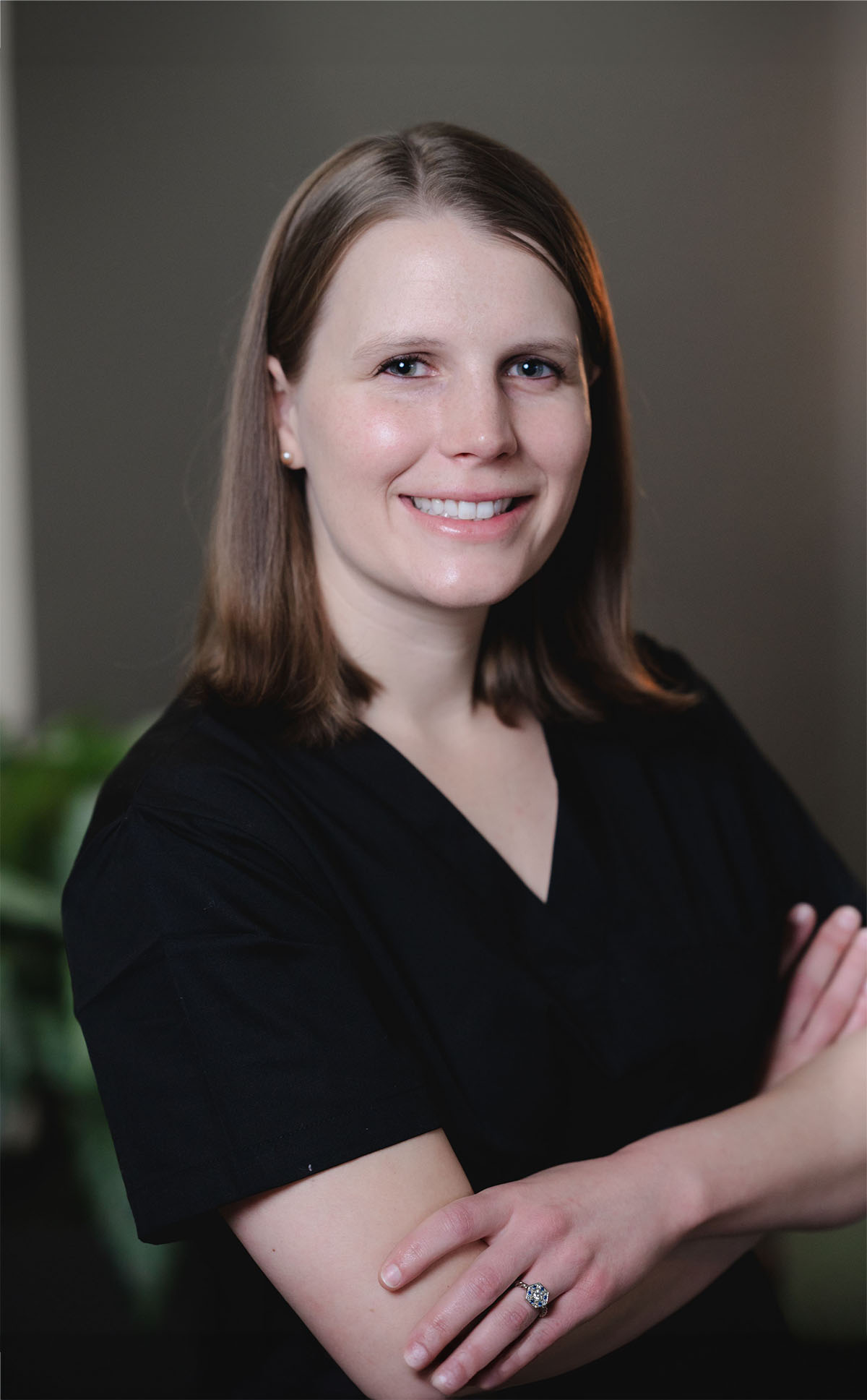 Smiling woman with shoulder-length light brown hair wearing black scrubs, arms crossed, and a ring on her finger.