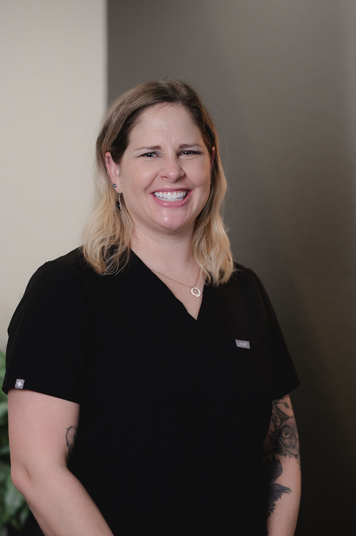 Smiling woman with blonde hair wearing black medical scrubs and visible tattoos on her arms.