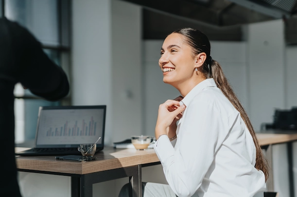 Smiling woman with long hair tied back sitting at a desk with a laptop displaying graphs.