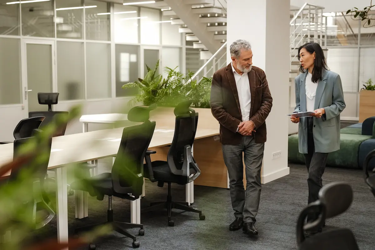 Two colleagues in business attire walking and talking in a modern office with plants and ergonomic chairs.