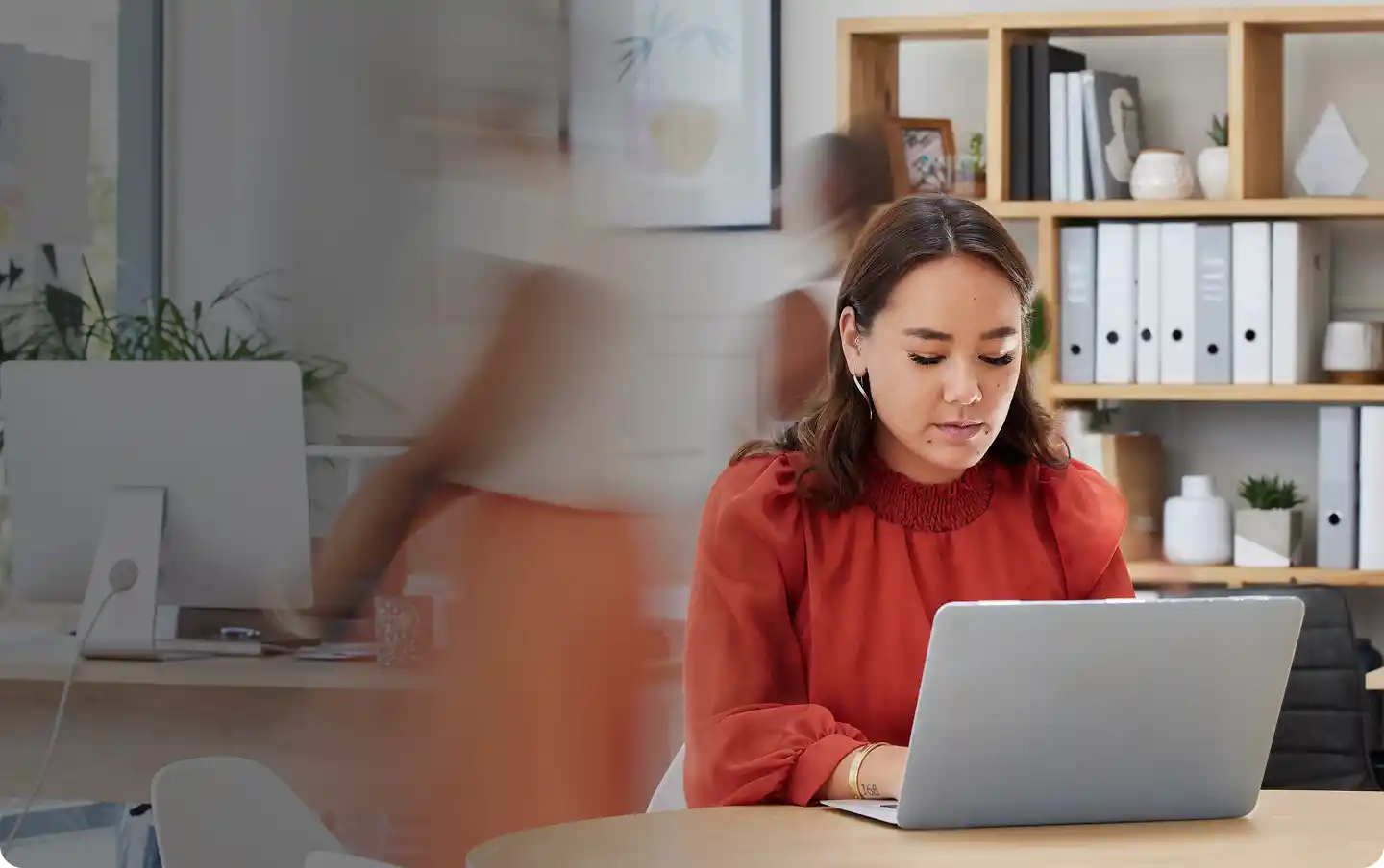 Une femme en chemisier rouge travaillant sur un ordinateur portable à une table ronde dans un bureau, avec des silhouettes floues qui se déplacent en arrière-plan.