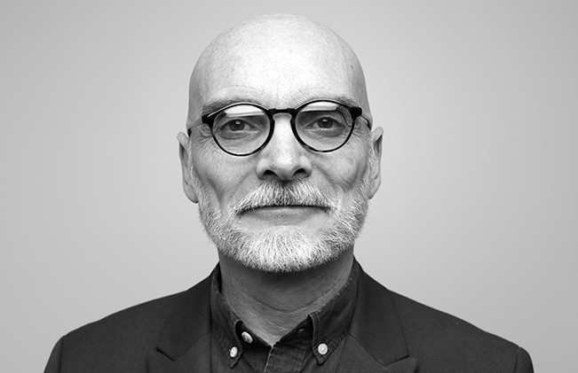 Black and white portrait of a bald man with glasses and a grey beard wearing a dark jacket and shirt.