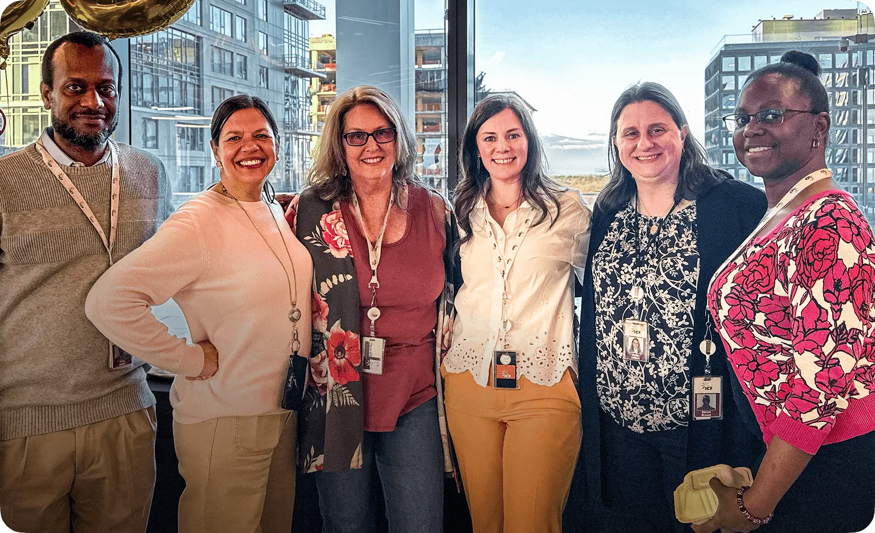 Six diverse coworkers smiling and posing together indoors with city buildings visible through the window background.