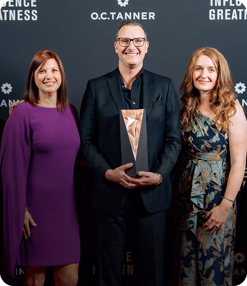 Three people standing and smiling at an award event, with the man in the center holding a geometric trophy.