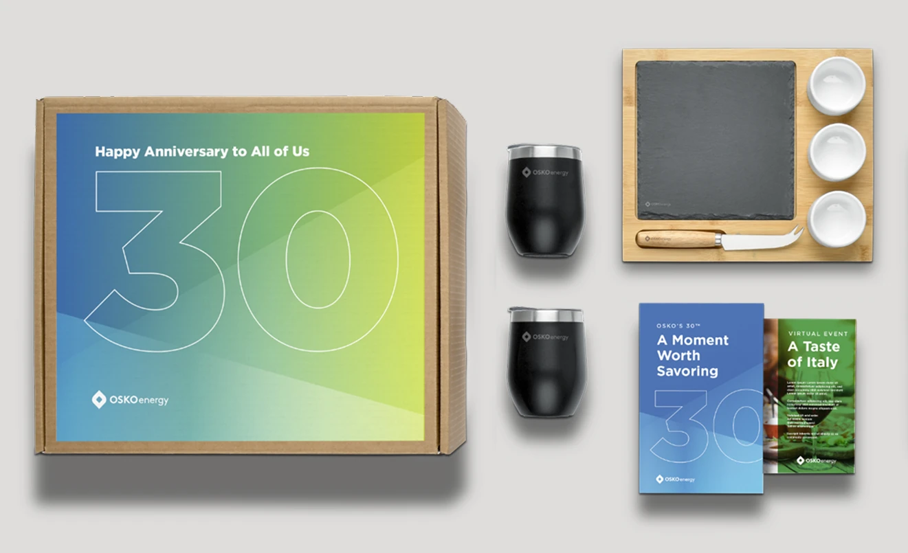 Coffret cadeau anniversaire comprenant deux verres OSKO Energy noirs, un plateau à fromage en bois avec ardoise et couverts, ainsi que des brochures intitulées « Un moment à savourer » et « Un avant-goût de l'Italie ».