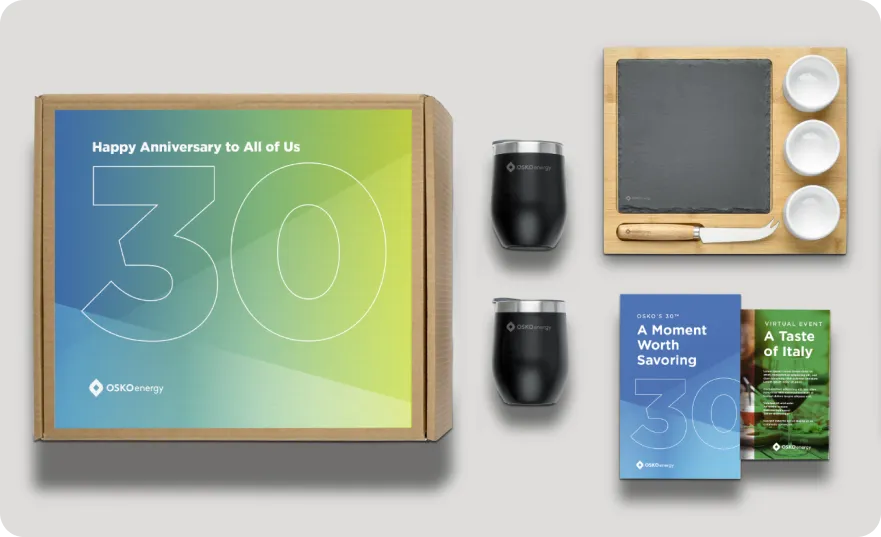 Anniversary gift box set including two black OSKO Energy tumblers, a wooden cheese board with slate and utensils, and event brochures titled 'A Moment Worth Savoring' and 'A Taste of Italy'.