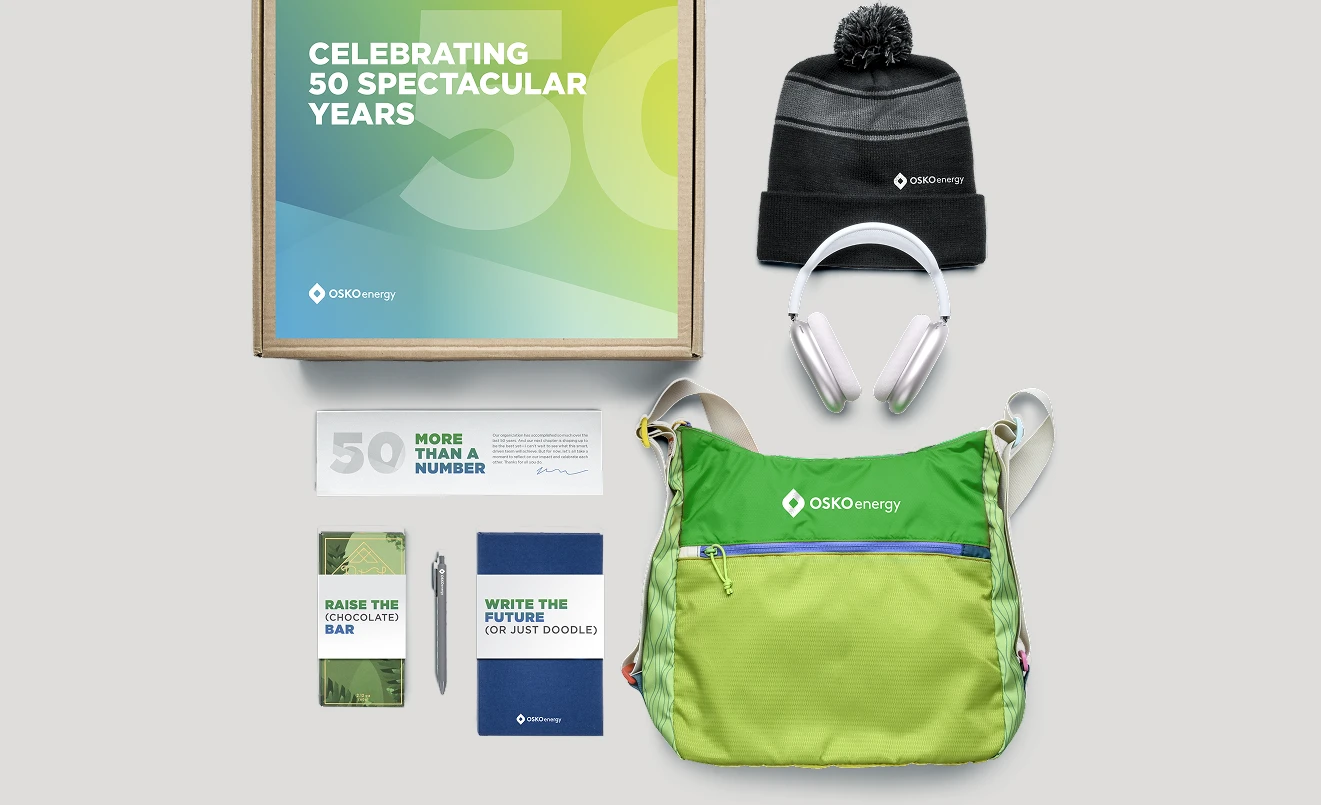 Collection of OSKO Energy branded items including a framed print celebrating 50 years, a black and gray beanie, white headphones, a green shoulder bag, a chocolate bar, a pen, and a notebook.