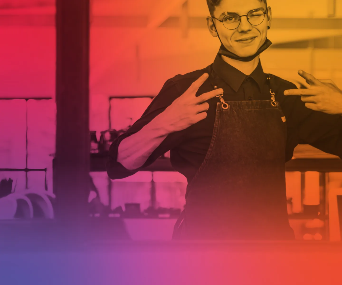 Young person wearing glasses and apron winking and showing peace signs with both hands in a colorful tinted restaurant or cafe setting.