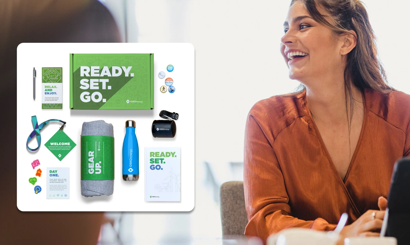 Smiling woman in brown shirt with a white overlay showing a green boxed gift kit including a blanket, water bottle, badge buttons, lanyard, mug, welcome card, pen, sunglasses, snack bar, and a notebook with 'READY. SET. GO.' text.
