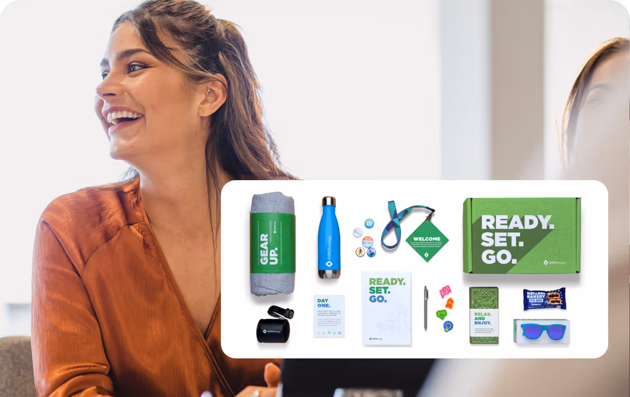 Smiling woman in brown shirt with a white overlay showing a green boxed gift kit including a blanket, water bottle, badge buttons, lanyard, mug, welcome card, pen, sunglasses, snack bar, and a notebook with 'READY. SET. GO.' text.