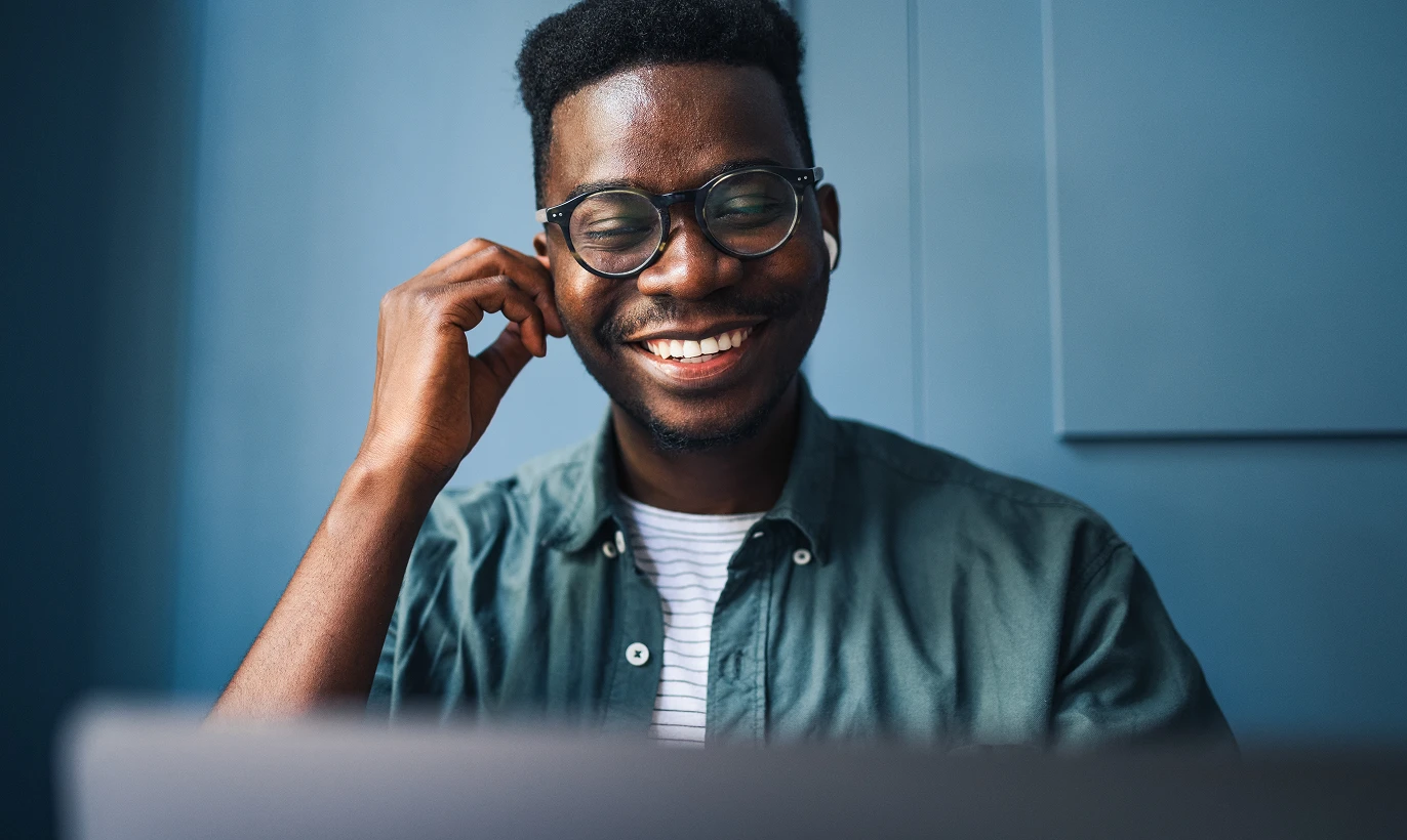 Smiling young man wearing glasses and earbuds, sitting in front of a laptop with a blue background.