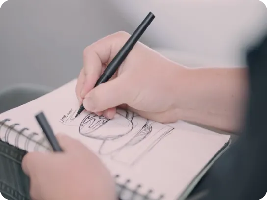 Person sketching a cartoon character on a spiral-bound notebook with a black pen.