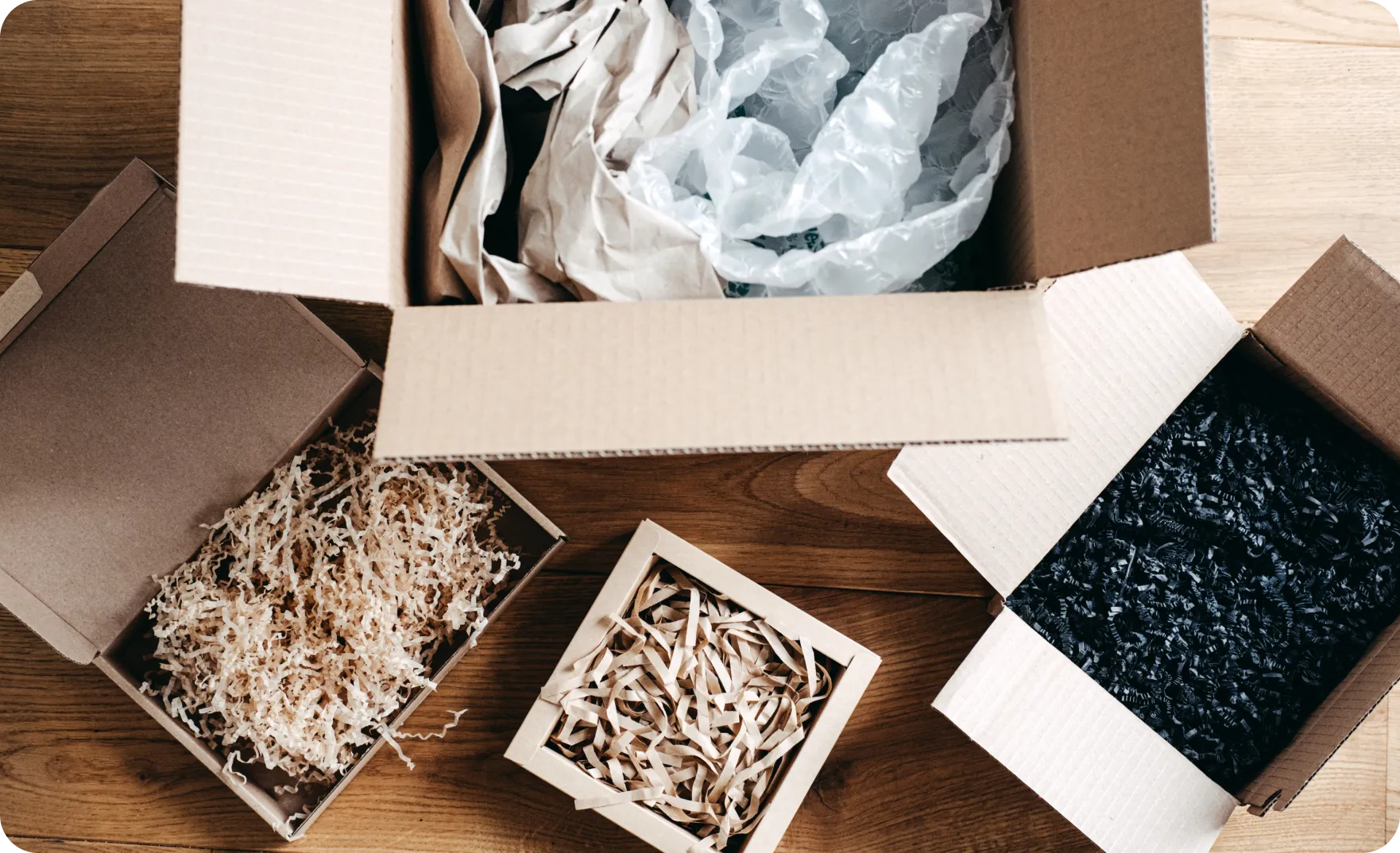 Open cardboard boxes filled with various packing materials including shredded paper, crumpled brown paper, bubble wrap, and black crinkle paper on a wooden surface.