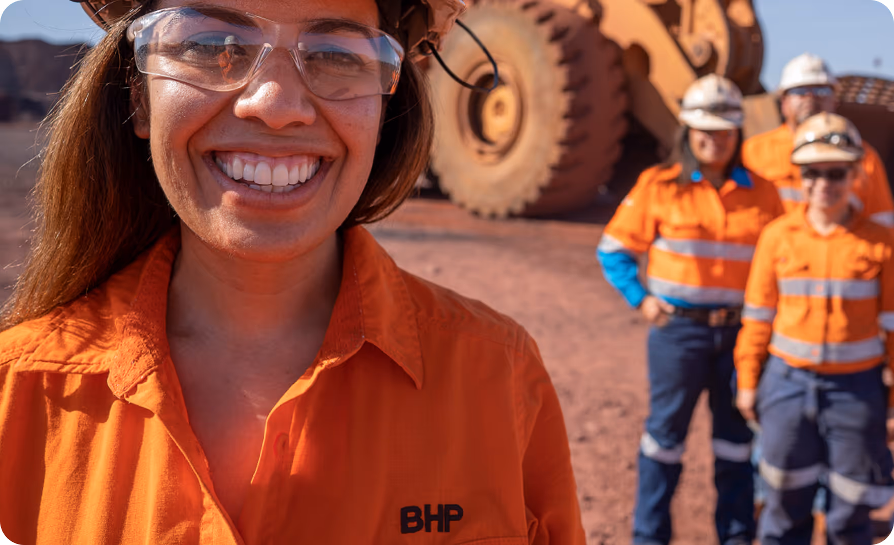 Smiling woman in orange work shirt and safety glasses with BHP logo, standing in front of three other workers in similar safety gear and a large mining vehicle.