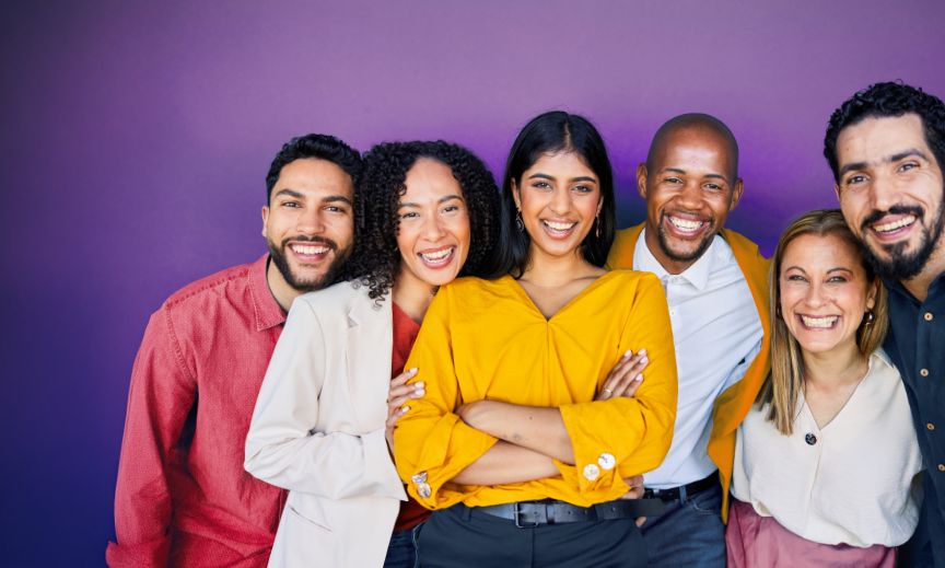 Diverse group of six smiling adults standing closely together in front of a purple background.