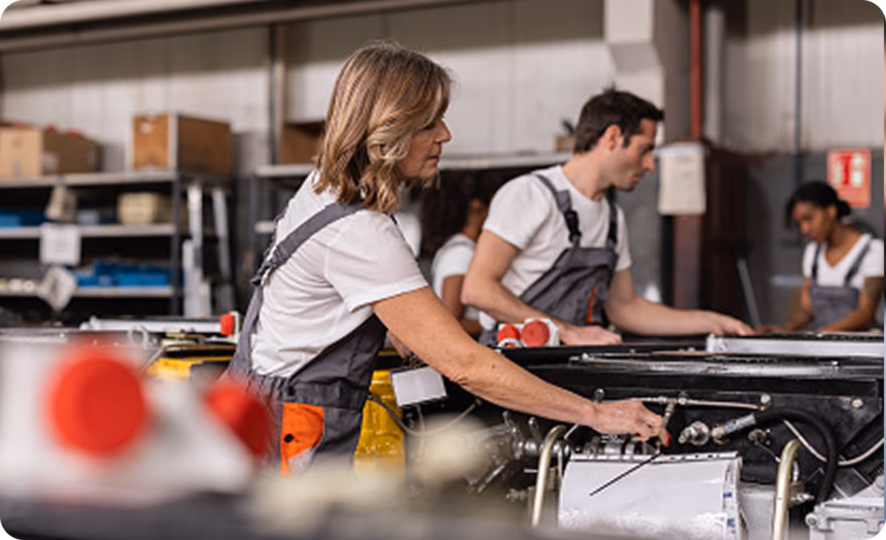 Workers in gray overalls operating and inspecting machinery in an industrial workshop.