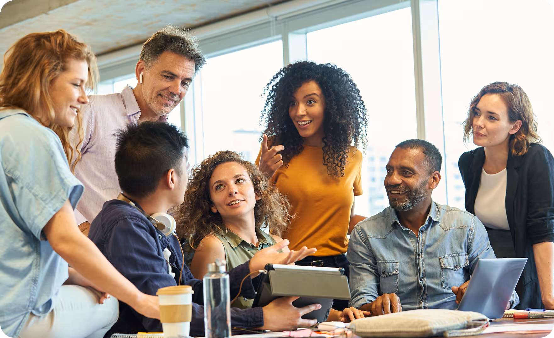Diverse group of six coworkers engaged in a lively discussion around a table with laptops and notebooks in a bright office.