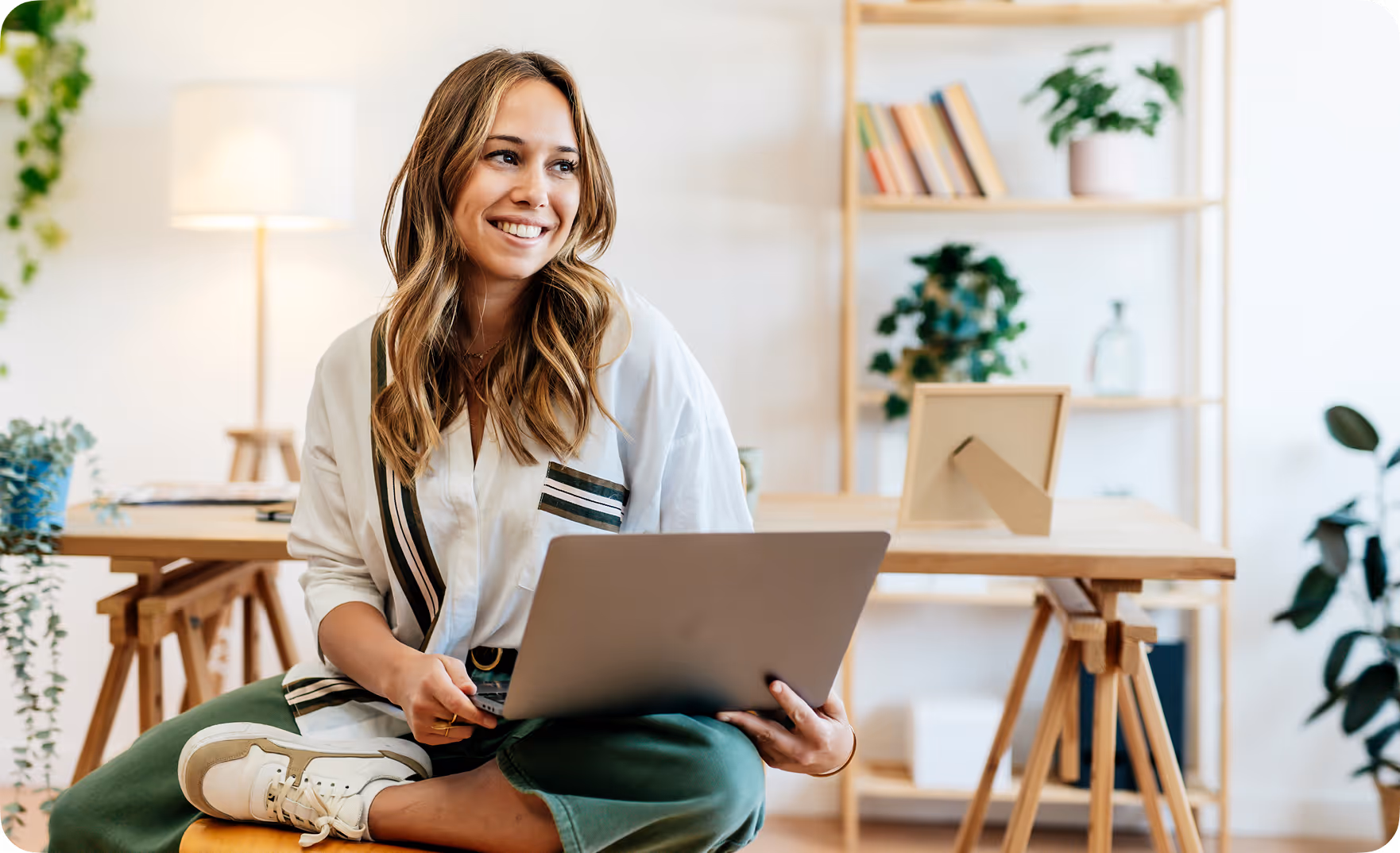 Smiling woman sitting cross-legged on a stool using a laptop in a bright home office with wooden furniture and plants.