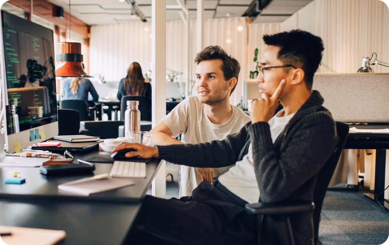 Two young men collaborating on a computer in a modern office setting with coding visible on the screen.