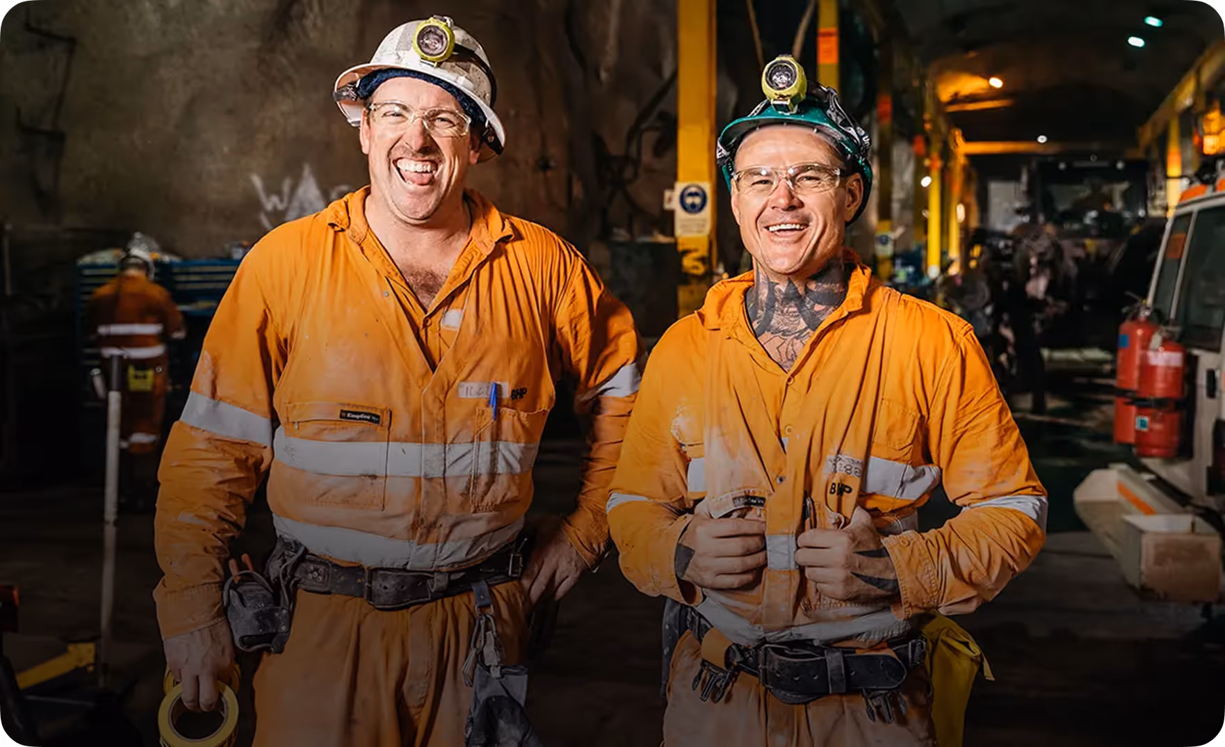 Two miners in orange high-visibility workwear and helmets with headlamps smiling inside an underground mine.