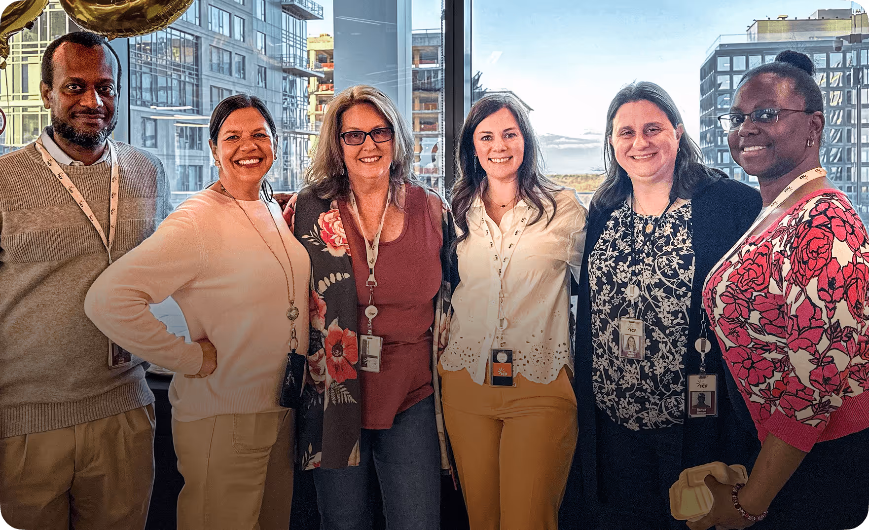 Group of six diverse professionals standing and smiling indoors with large windows showing city buildings in the background.