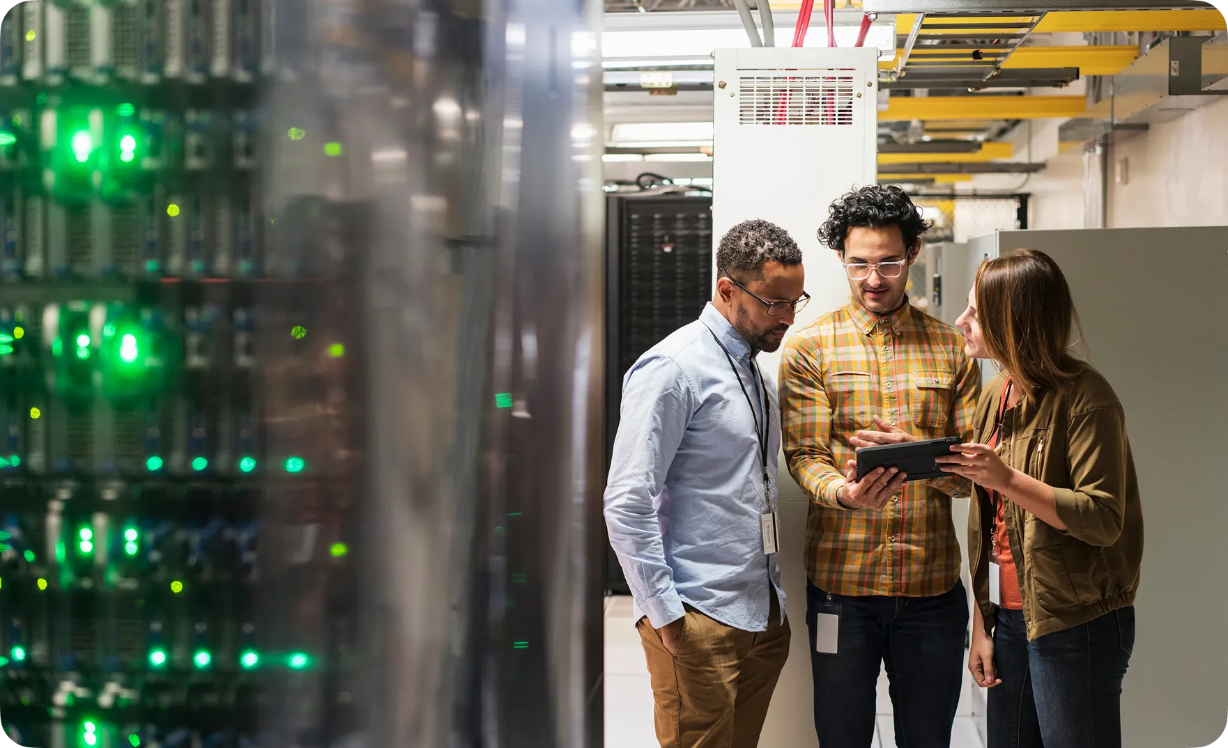 Three IT professionals discussing data on a tablet in a server room with racks and green indicator lights.