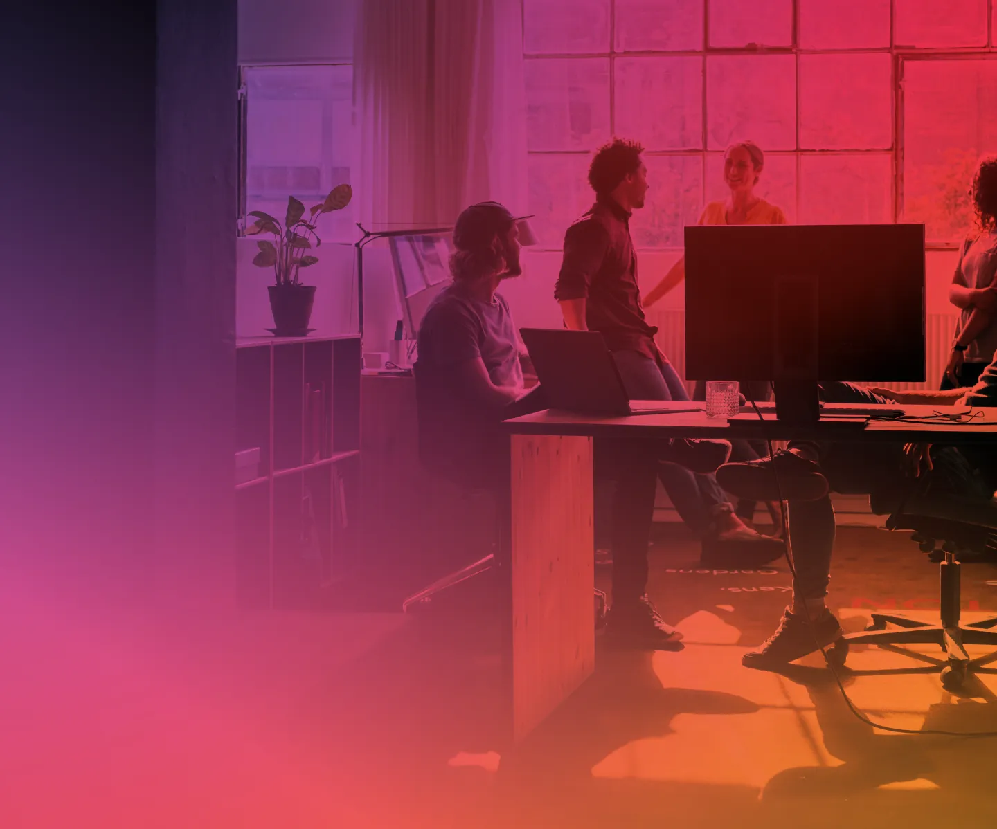 Group of young professionals engaging in a discussion in a modern office with large windows and a computer on the desk.