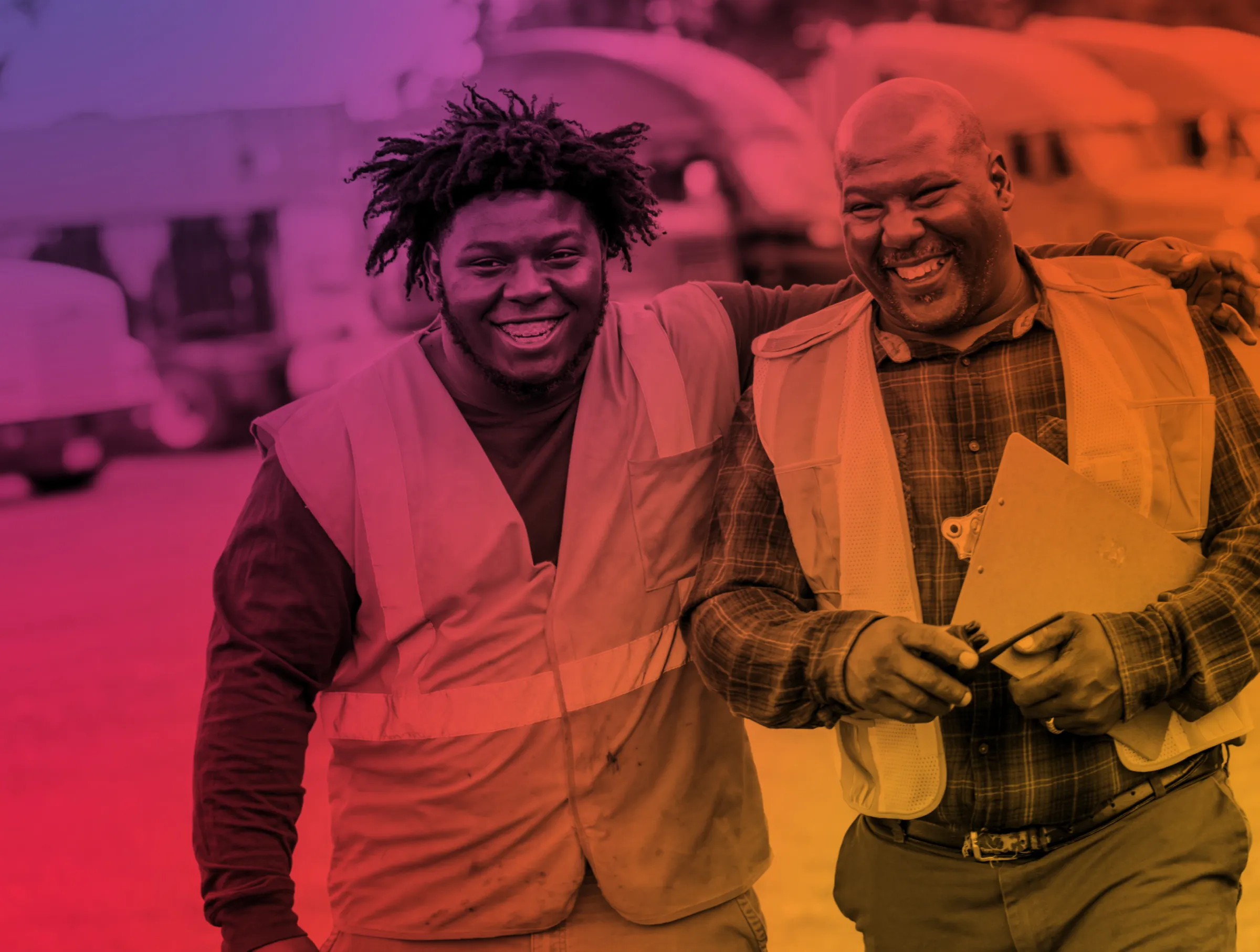 Two happy construction workers in safety vests smiling with one worker holding a clipboard and a pen.