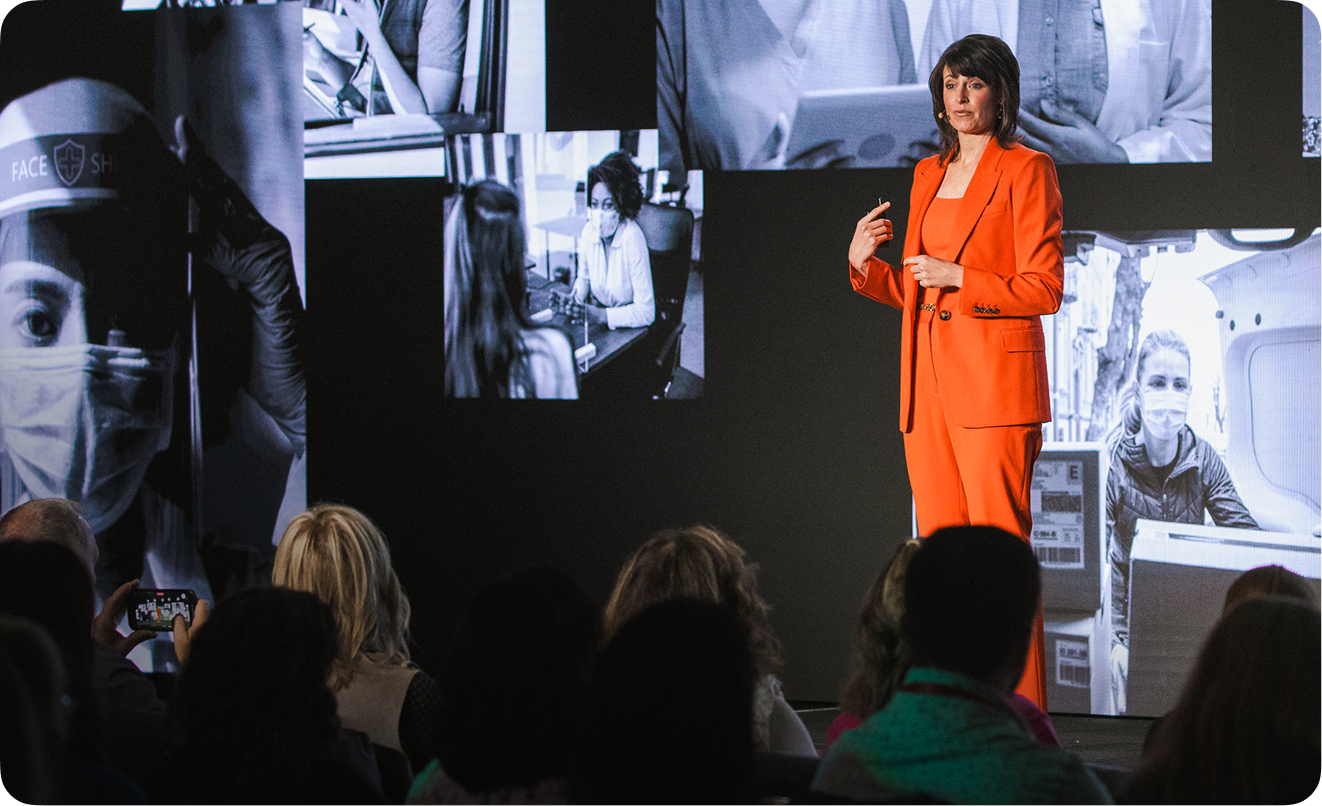 Woman in an orange suit speaking on stage to an audience with a backdrop showing images of people wearing masks and in meetings.