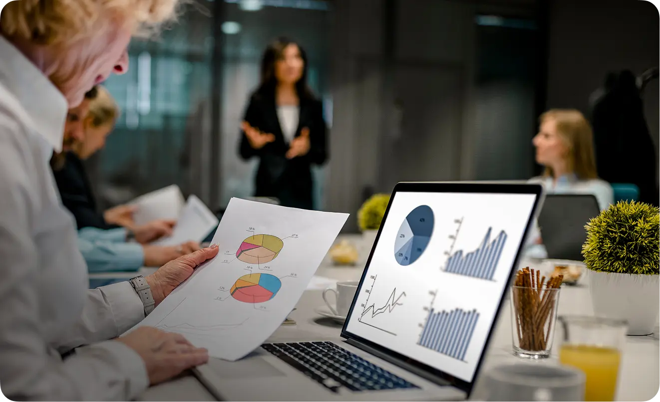 Person holding a paper with pie charts while viewing graphs and charts on a laptop screen during a business meeting.