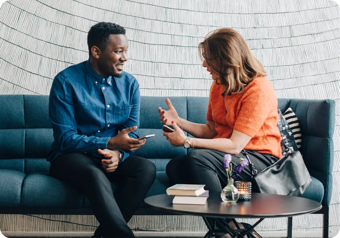 A man and woman sitting on a blue sofa having an engaged conversation while holding smartphones, with a black round table in front holding books, flowers in a vase, and a candle.