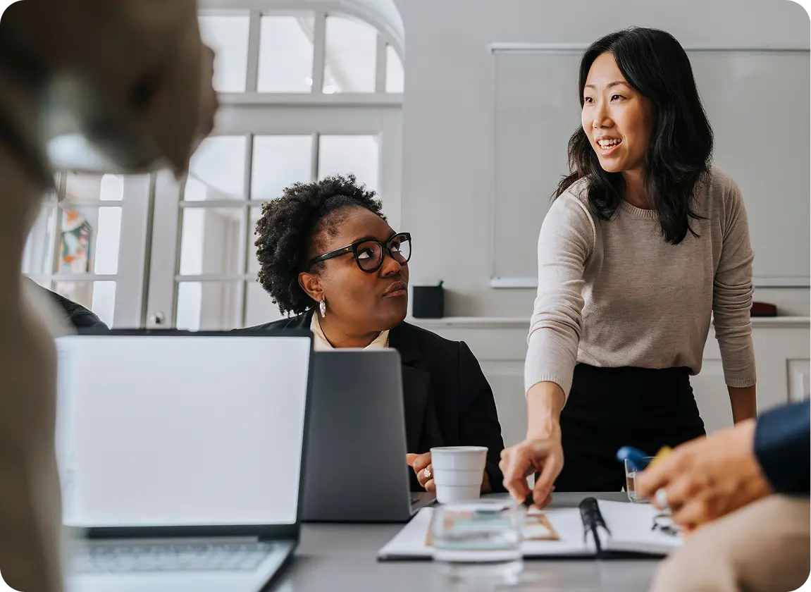 Two women collaborating in a bright office, one standing and smiling while pointing at documents on a table, the other seated and attentively listening with a laptop in front.