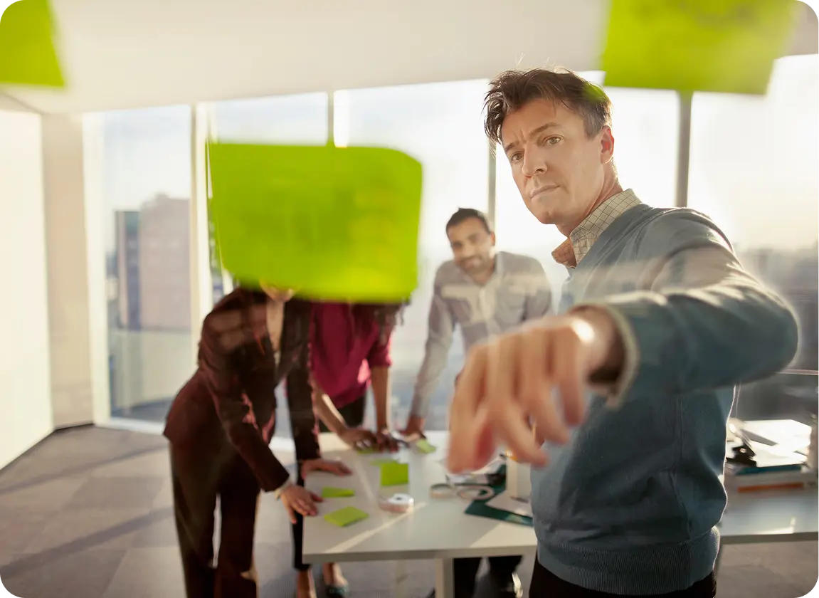 A man writing on a glass board with green sticky notes while three colleagues watch and discuss around a table in a bright office.