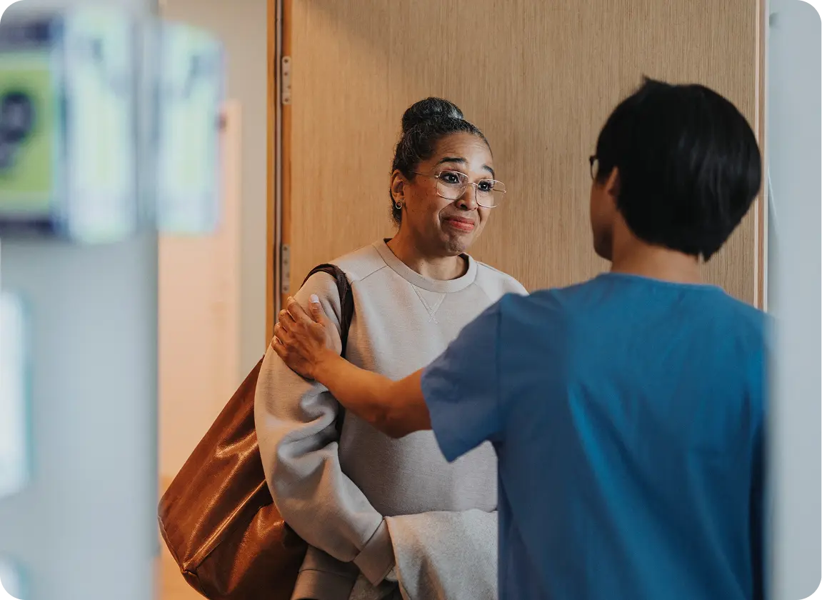 Healthcare professional in blue scrubs comforting a woman with glasses and a brown bag by gently holding her upper arm.
