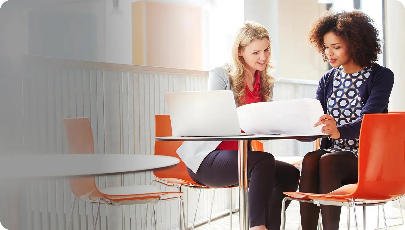 Two women sitting at a table with a laptop, reviewing large paper documents in a bright modern office.