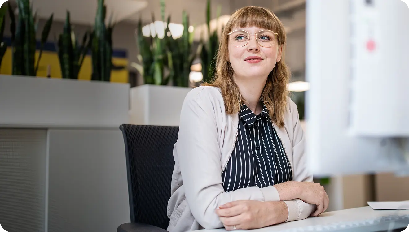 Young woman with glasses sitting at a desk, smiling and looking at a computer screen in a modern office.
