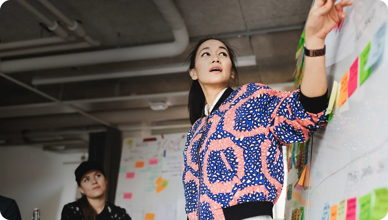 Woman in vibrant patterned jacket pointing to sticky notes on a wall during a meeting while another woman attentively watches.