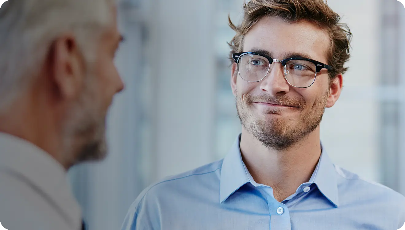 Young man with glasses smiling slightly while conversing with an older man in a professional setting.