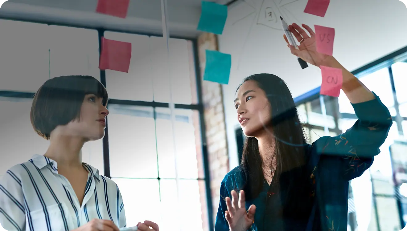 Two women collaborating in an office, discussing ideas with sticky notes on a glass wall.