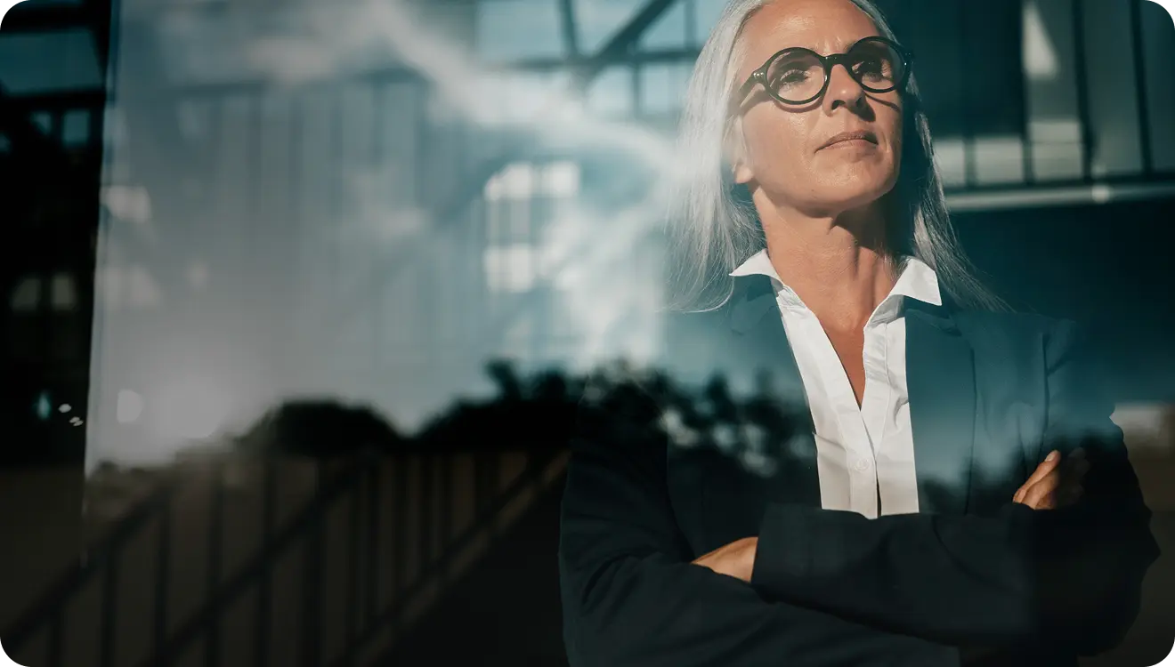 Confident older woman with grey hair and glasses, arms crossed, standing indoors with a reflective glass window showing an architectural structure.
