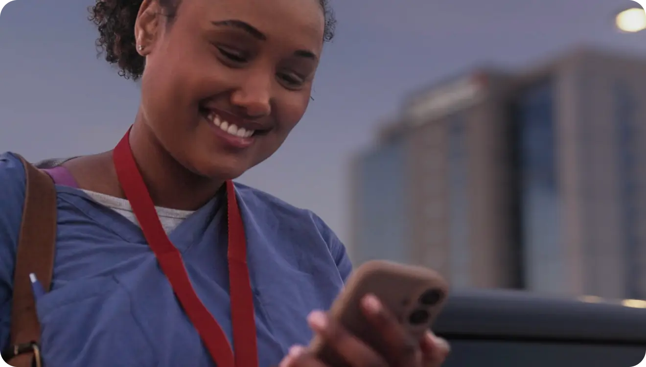 Smiling woman in a blue top looking at her smartphone outdoors near a building at dusk.