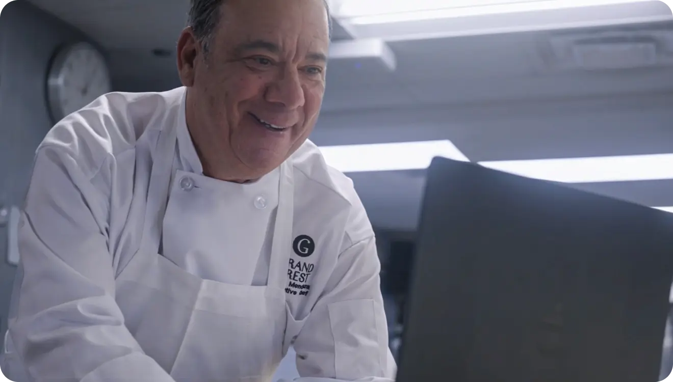 Smiling male chef in white uniform looking at a laptop in a kitchen.