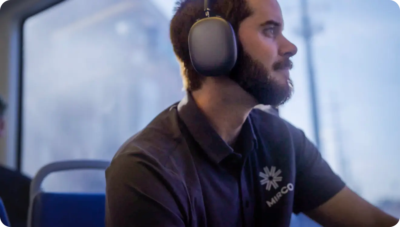Man wearing large noise-canceling headphones and a black shirt with a MIRCO logo, sitting on a bus or train.