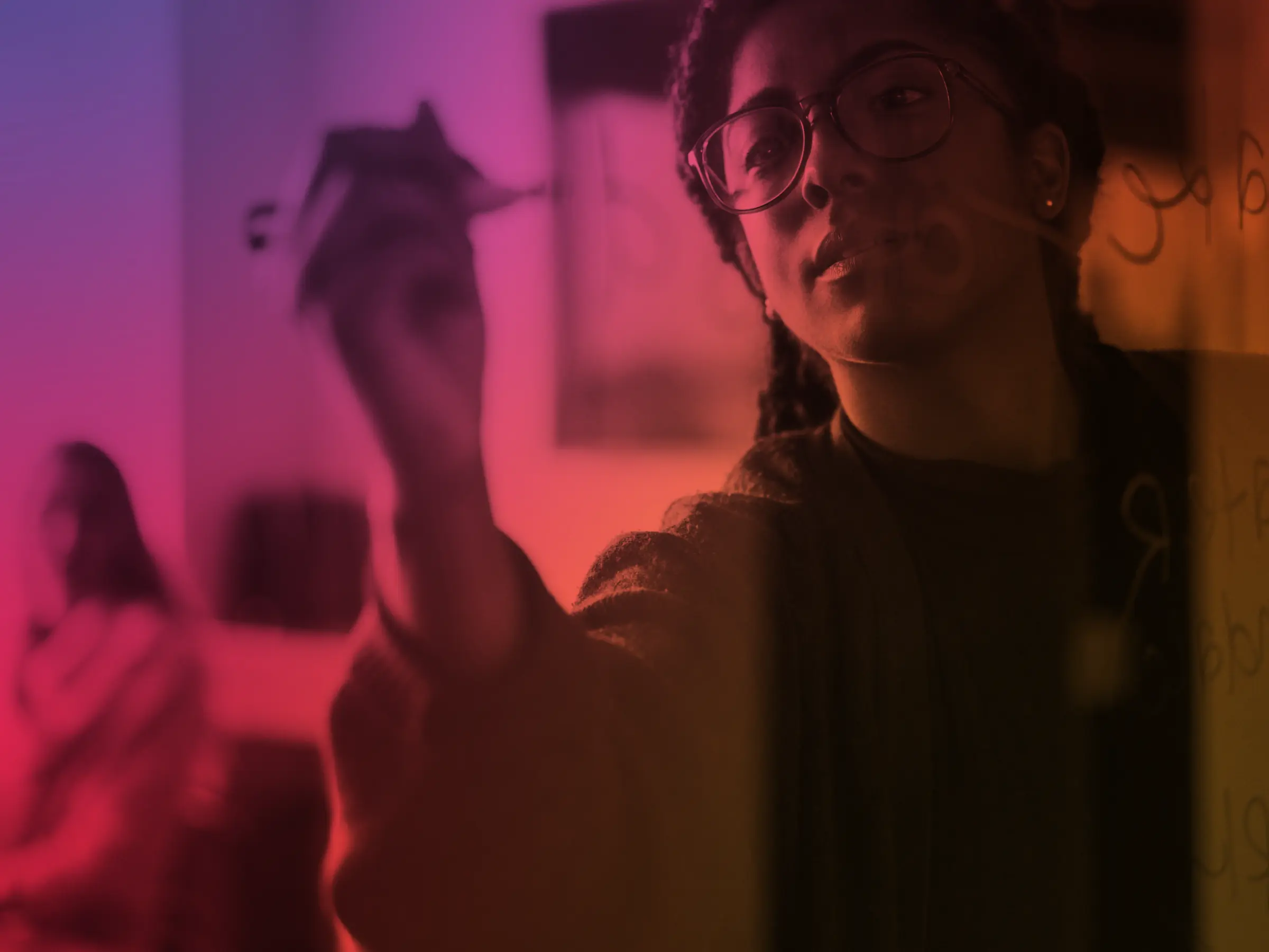 Person with glasses writing on a transparent board with another person blurred in the background.