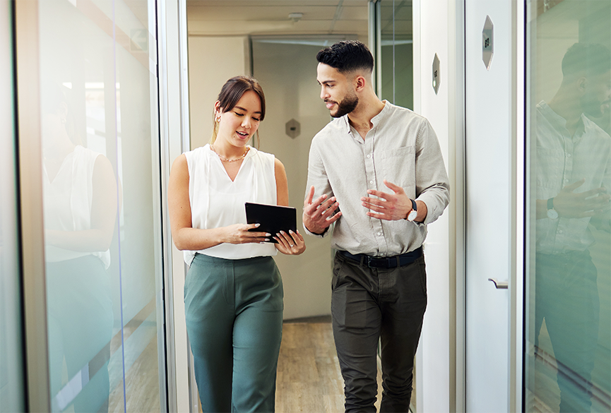 Man and woman in business attire, walking and talking down a hallway with glass windows holding a tablet between the two of them.