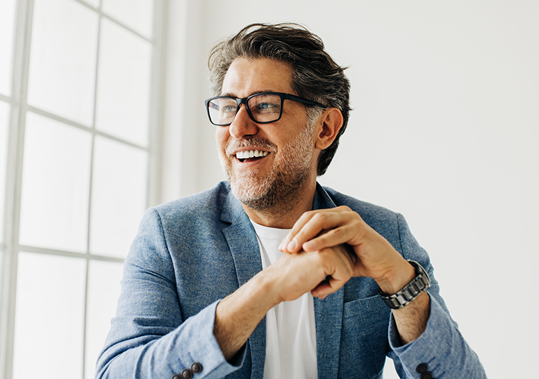A zoomed in image of a middle aged man in a bright room with a suit on, sitting down with his arms in front of him.