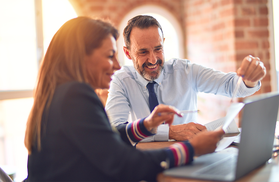  A woman in a suit jacket sitting in front of a laptop and holding a piece of paper with a man in a button down shirt and tie across from her, pointing to the laptop screen with a pen.
