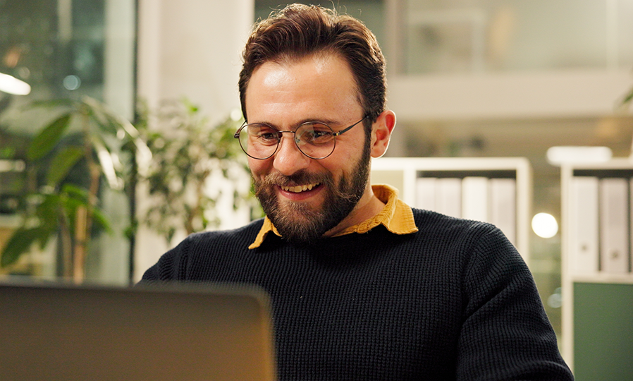 A man in glasses with a sweater on sitting at a desk, smiling at a laptop screen.