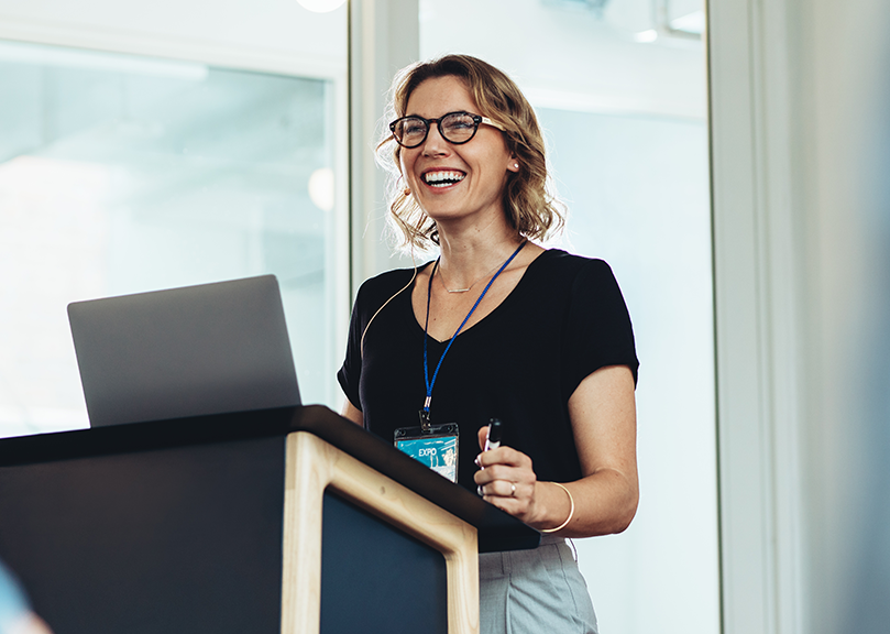 A professional woman standing at a podium, smiling at an audience out of frame. 